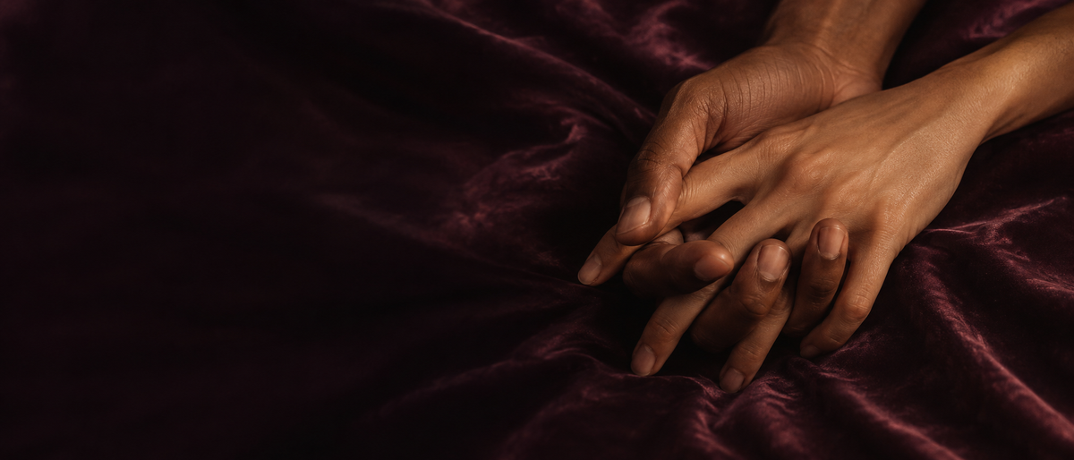 Two hands loosely interlaced on dark plum velvet in warm low light